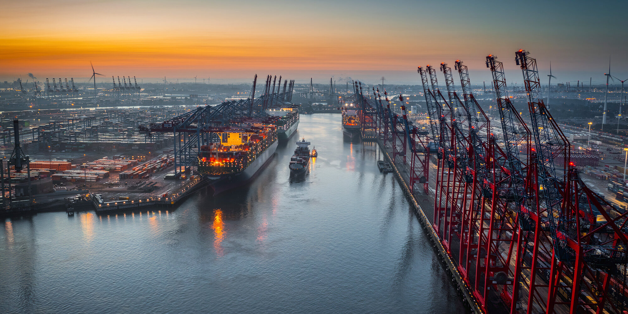 Aerial view of a container terminal with large container cargo ships and gantry cranes. A cargo ship  is navigating through the central waterway.  On both sides of the waterway are massive container terminals lined with red and black gantry cranes used for loading and unloading ships.  on the left docked ships with brightly lit containers. Moody sky at dawn. Port of Hamburg in Germany, Europe.