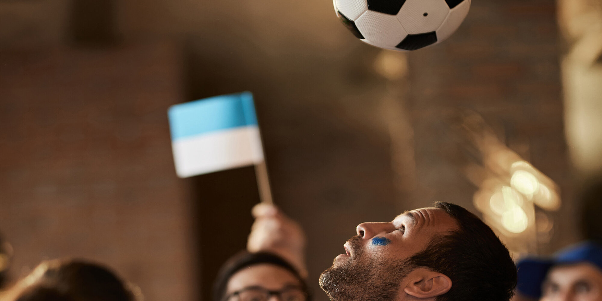 Mann in Fussballtrickot köpfelt einen Fussball nach oben, hinten jubeln Fans