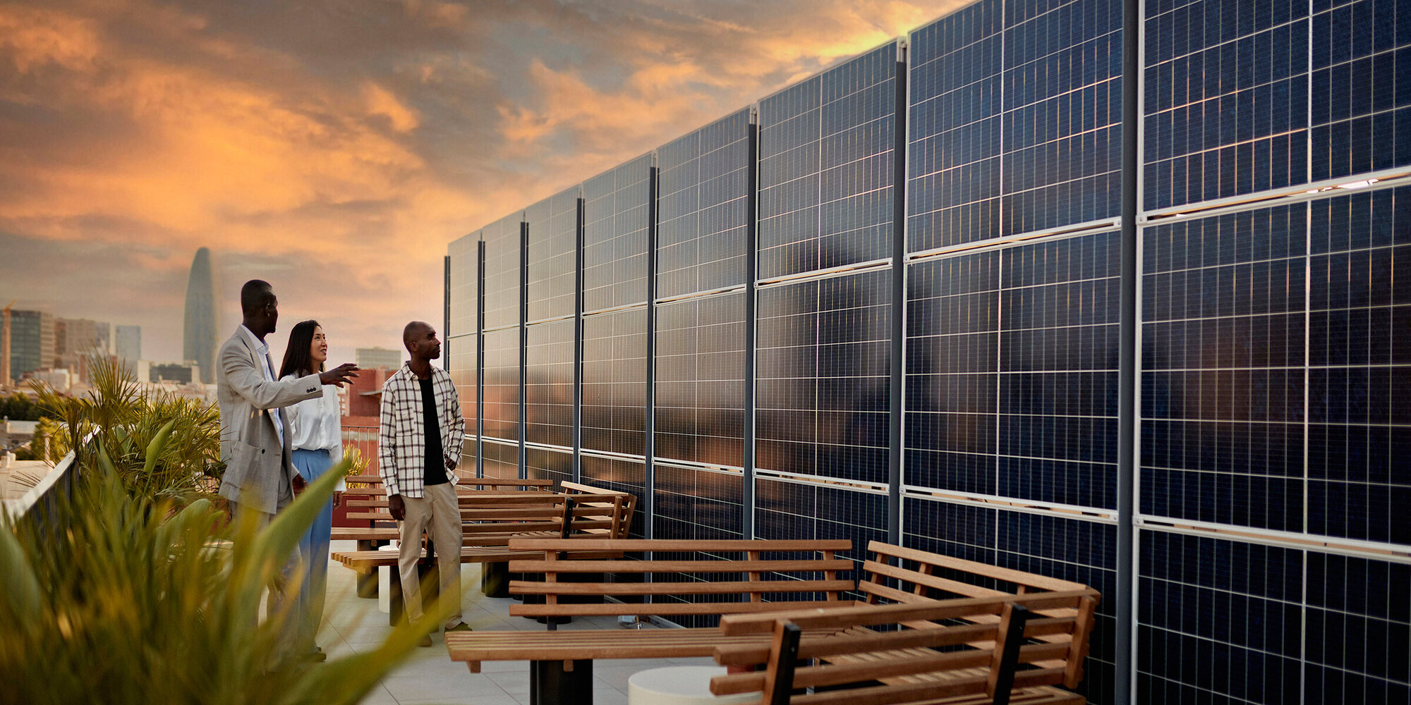 Real estate agent and couple standing on rooftop of environmentally aware office building with dramatic sky and Barcelona cityscape in background at sunset.