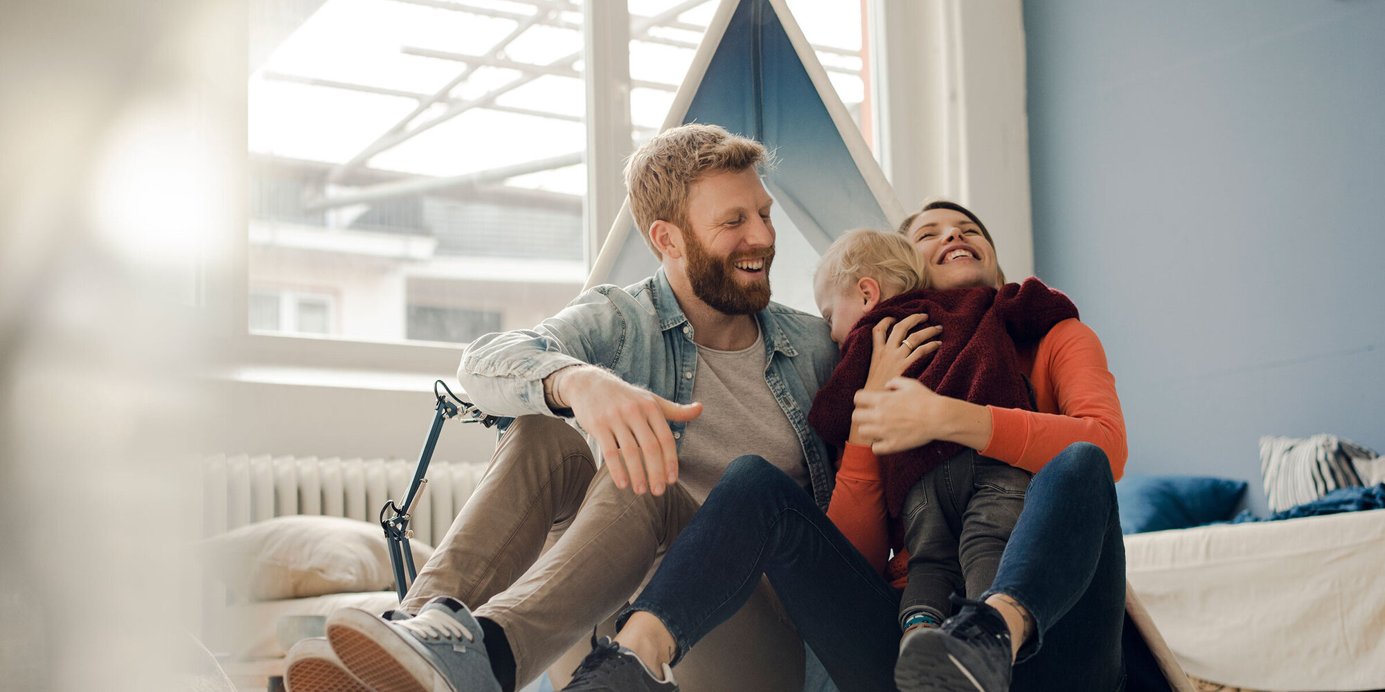 Familie mit Kleinkind sitzt im Kinderzimmer am Boden und umarmen das Kind