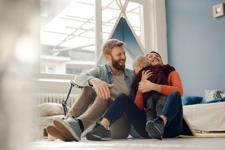 Familie mit Kleinkind sitzt im Kinderzimmer am Boden und umarmen das Kind