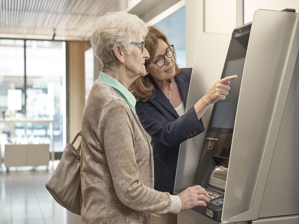 Eine Kundin wird am Bancomaten von einer Mitarbeitenden unterstützt.
