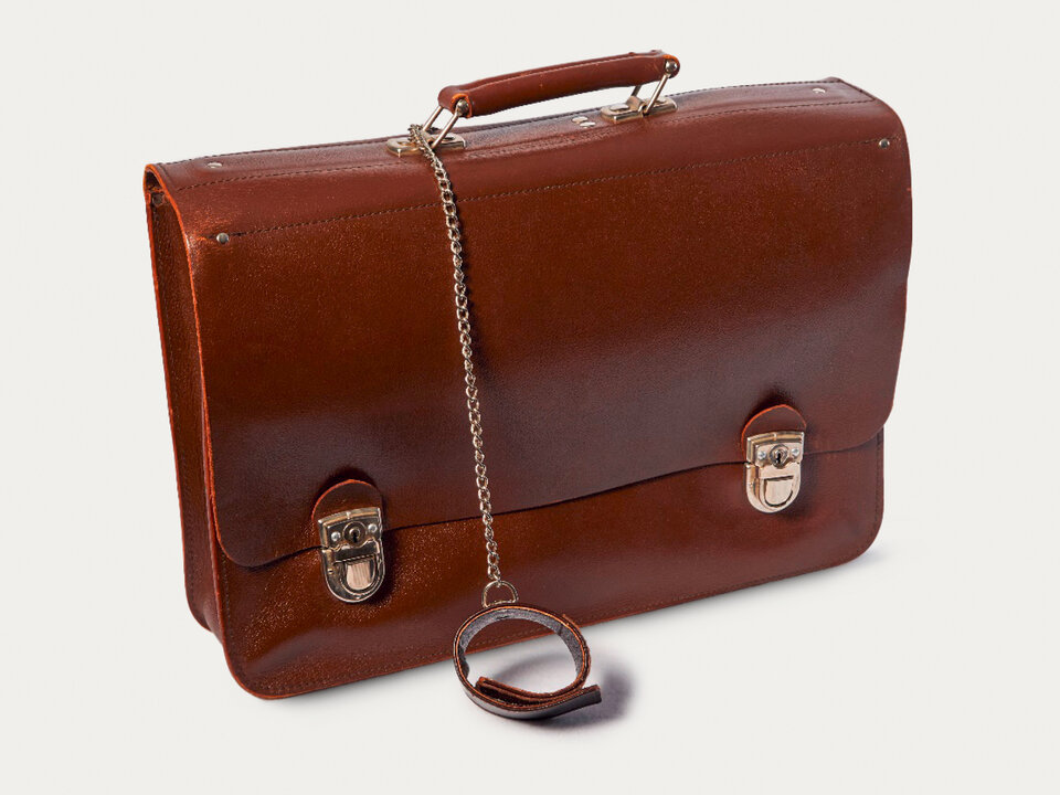 Das Bild zeigt eine ältere lederne Tasche, an deren Handgriff eine Kette mit einem Klettverschlussband angebracht ist.