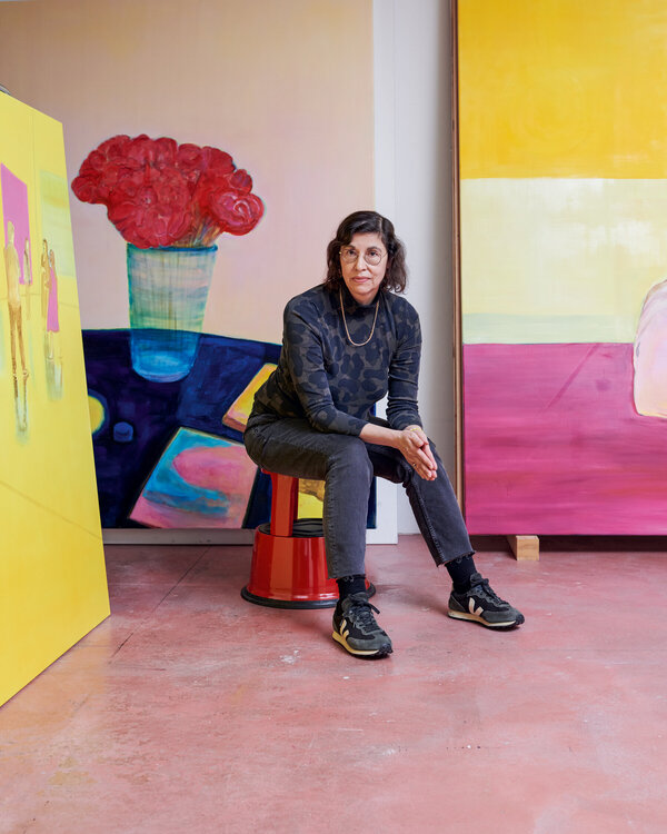 Das Bild zeigt Künstlerin Caro Niederer, wie sie in ihrem Zürcher Atlier auf einem Hocker sitzt, umringt von Gemälden.